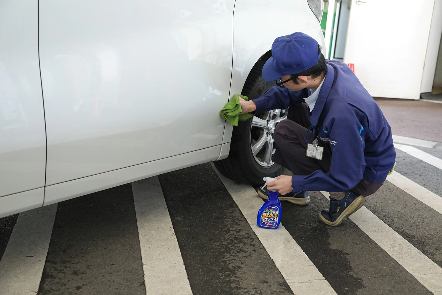 作業員が白い車の側面を洗浄用スプレーと布で丁寧に磨いている様子。制服と名札を着用し、整備エリアで作業中。